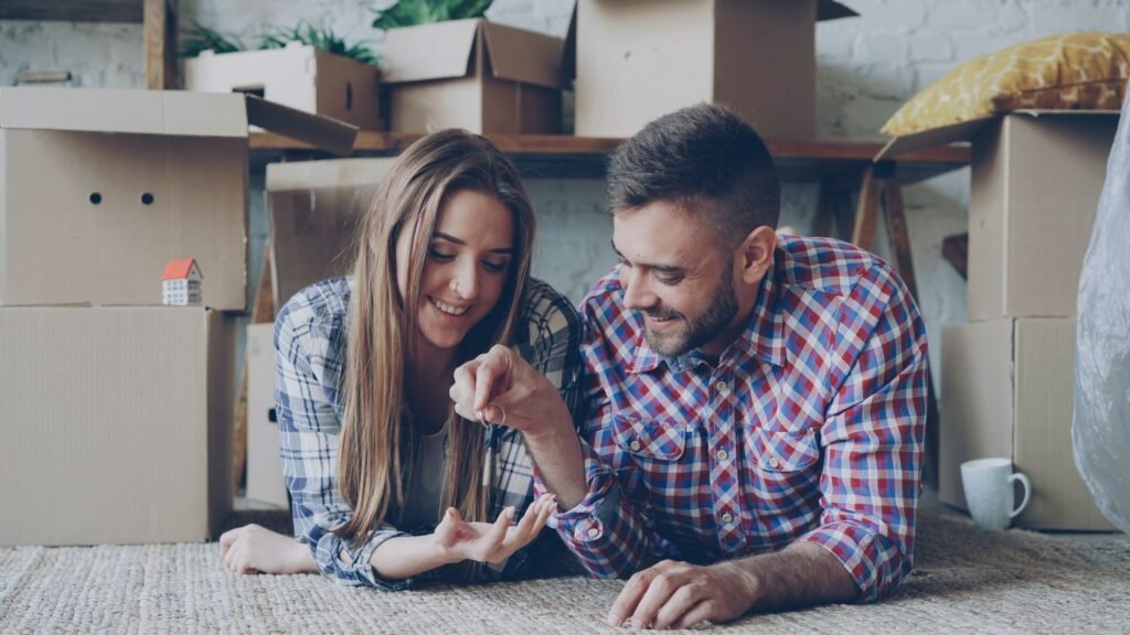 Couple sitting on the floor amid moving boxes smiling and holding house keys after buying their first home in Dubai