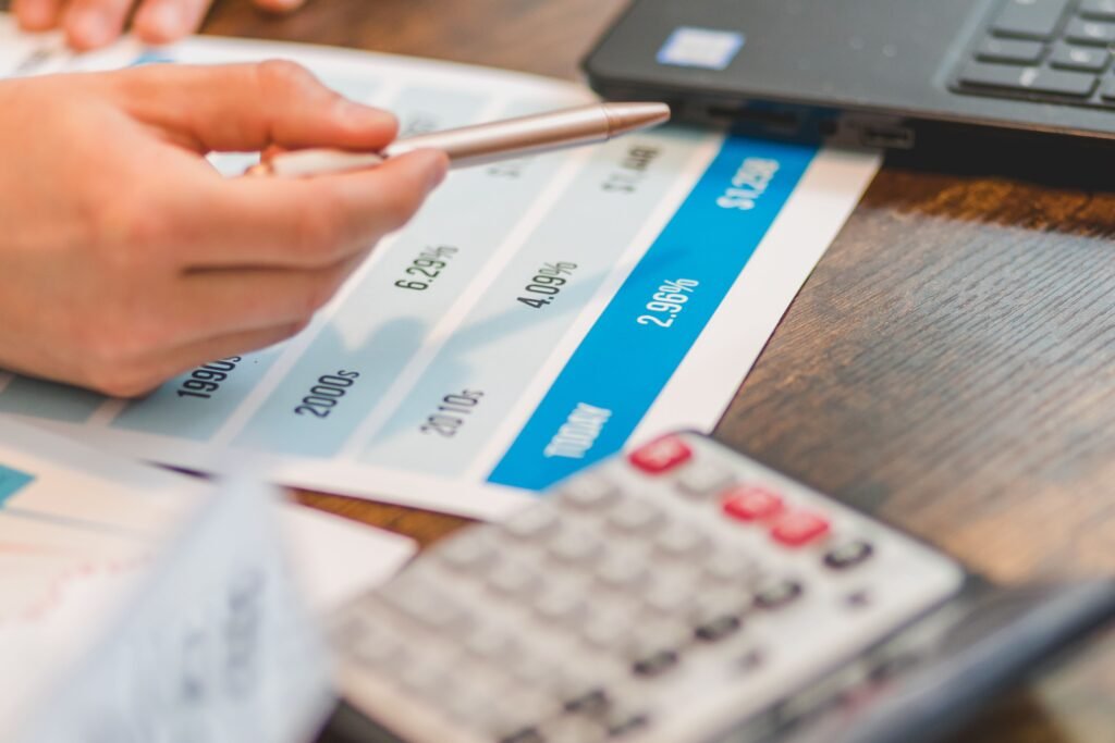 Hand pointing a pen at a printed chart showing rising interest rate percentages next to a laptop and calculator
