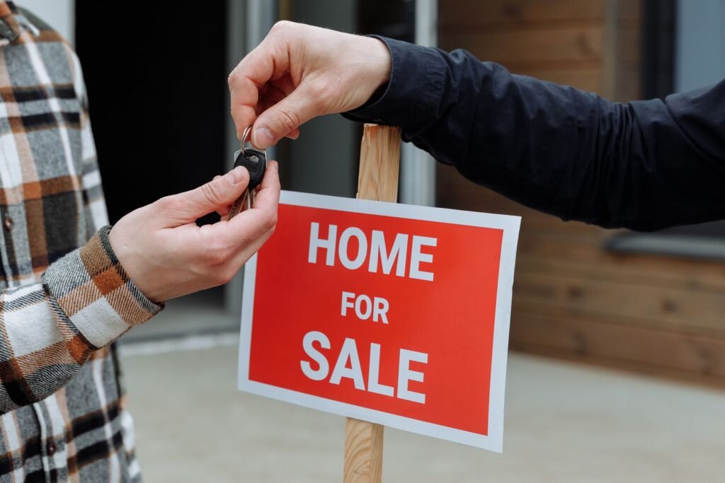 Hands passing a set of keys beside a Home For Sale sign, the handover moment when a mortgaged property is sold and the bank lien is released